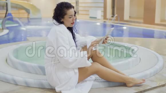 浴袍上的美女在游泳池附近使用数码平板电脑视频的预览图