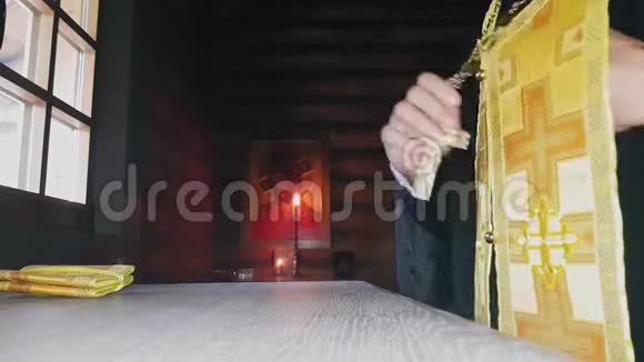 正教的牧师在暮色中穿着神圣的衣服站在木教堂祭坛的窗户前视频的预览图
