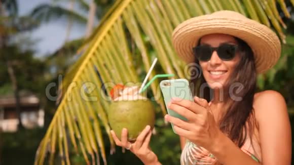 年轻快乐的混血旅游女孩在手机上发短信在热带海滩喝椰子鸡尾酒4公里视频的预览图