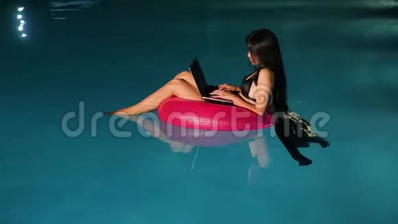 年轻漂亮的黑发女人在游泳池里用笔记本电脑工作视频的预览图