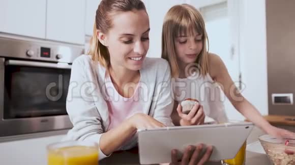 漂亮的年轻母亲和女儿在家厨房吃早餐时使用数码平板电脑视频的预览图