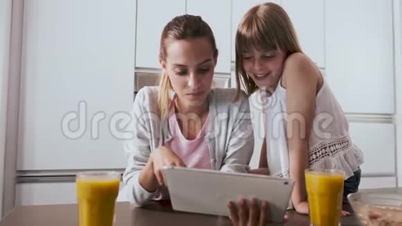 漂亮的年轻母亲和女儿在家厨房吃早餐时使用数码平板电脑视频的预览图