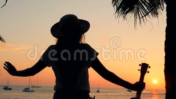 在美丽的热带海滩日落时手持Ukulele吉他站在伸展的地方无忧无虑的年轻少女视频的预览图