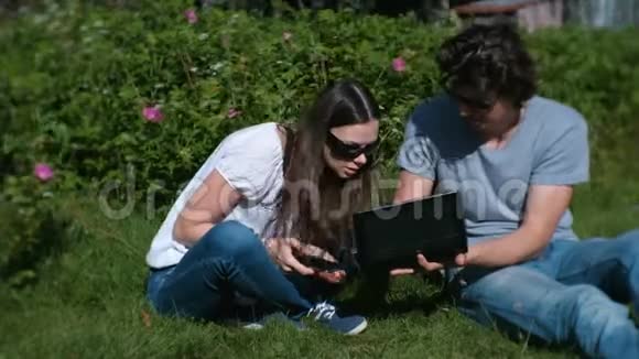 年轻夫妇一男一女坐在公园里一起用手机和笔记本电脑工作视频的预览图