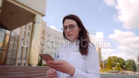 商业女性穿着白色衬衫在城市街道上行走和使用智能手机的正面视图慢镜头视频的预览图