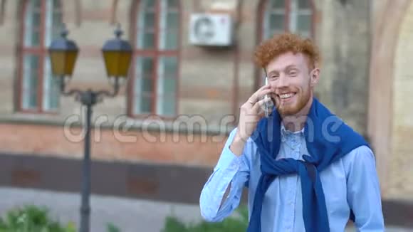 幸福的微笑男人留着姜黄色卷发和胡须在模糊的背景下通过手机交谈视频的预览图