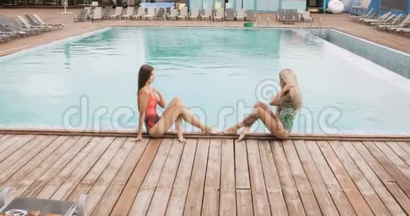 两个女孩在酒店泳池边摆姿势年轻女子穿着一件泳衣坐在游泳池的边缘摆姿势视频的预览图
