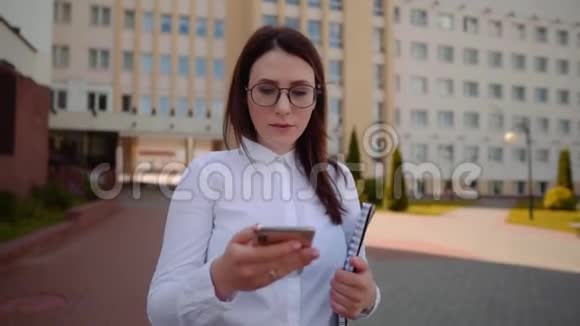 商业女性穿着白色衬衫在城市街道上行走和使用智能手机的正面视图慢动作镜头视频的预览图