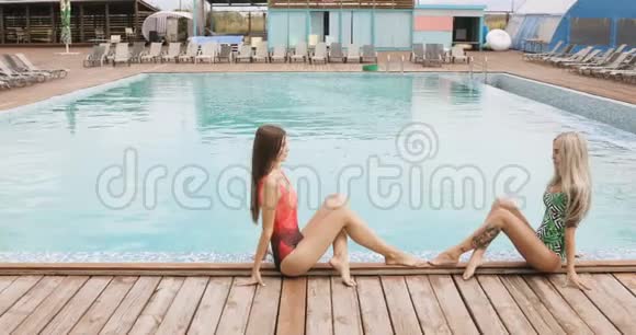 两个女孩在酒店泳池边摆姿势年轻女子穿着一件泳衣坐在游泳池的边缘摆姿势视频的预览图