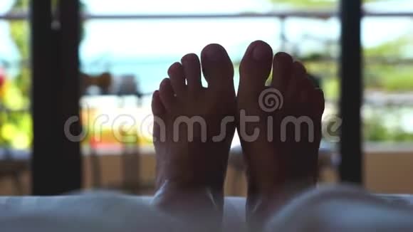 女人躺在旅馆的床上靠近挥动的双脚与热带海景3840x2160视频的预览图