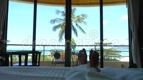 穿着浴袍的女人躺在床上挥舞着她的脚对着热带海景3840x2160视频的预览图