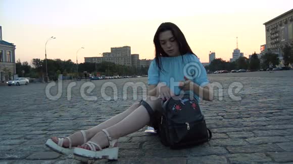 年轻漂亮的黑发女人坐在城市的道路上把耳机放在背包里视频的预览图