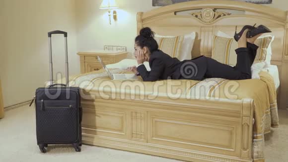漂亮的红唇女商人躺在酒店房间的床上使用笔记本电脑视频的预览图