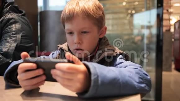 小男孩正在咖啡馆里玩电话带着电话的金发男孩视频的预览图