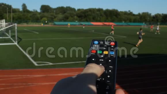 在足球场背景下一只手握着电视机遥控器的雄性手在足球场上踢球视频的预览图