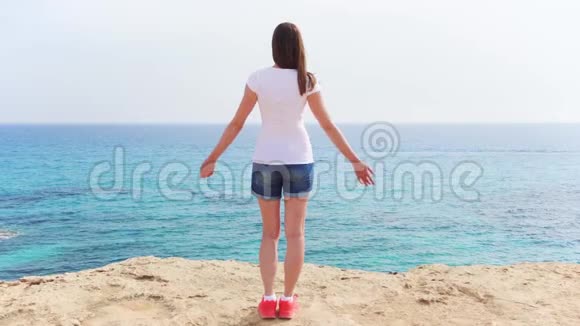 女人在碧海的峭壁上举起双臂女旅行者缓慢地伸出双手视频的预览图