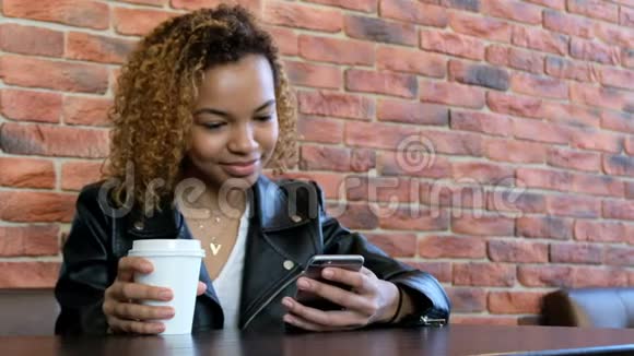 一个穿着皮夹克带着一杯白咖啡和手机的漂亮黑人女孩坐在桌子上视频的预览图