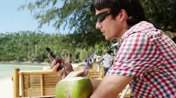 英俊的年轻商人戴着太阳镜在海滩咖啡馆的背景海景使用手机3840x2160视频的预览图