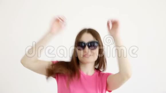 戴着太阳镜穿着粉色上衣和牛仔裤的漂亮快乐女孩在白墙上欢快地跳舞视频的预览图