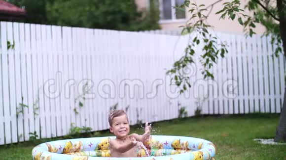 一个四岁的男孩躺在一个孩子充气游泳池里溅起水花微笑着附近的漂浮玩具在花园里在视频的预览图