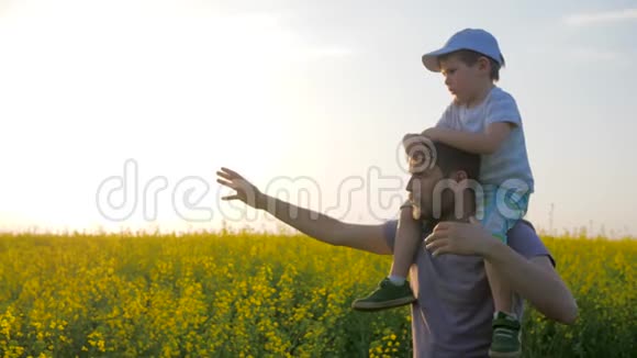 在阳光明媚的天气里爸爸和孩子在田野散步儿子在父亲的肩膀上花草地上的花孩子和爸爸在一起视频的预览图