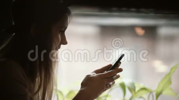 年轻时尚女性戴着帽子坐在窗户附近并使用智能手机的肖像女人用手机聊天视频的预览图