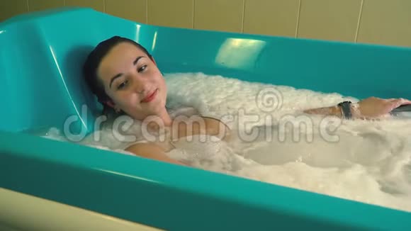 一个放松的女孩躺在泡沫浴缸里做水疗按摩手上拿着一个防水钟慢动作视频的预览图