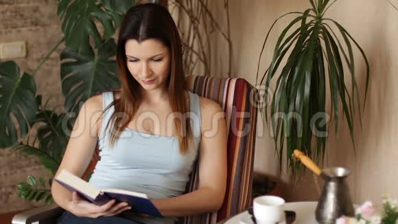 年轻迷人的女人在客厅里坐在舒适的椅子上读有趣的书关门视频的预览图