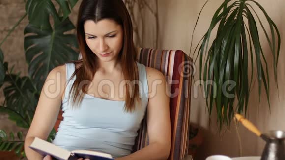年轻迷人的女人在客厅里坐在舒适的椅子上读有趣的书关门视频的预览图
