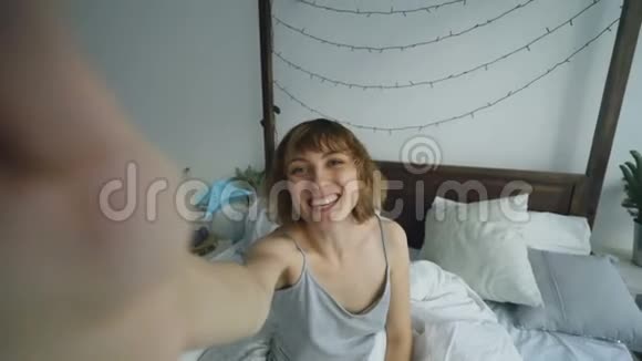 快乐的女人在家里坐在床上与朋友用智能手机摄像头在线视频聊天视频的预览图