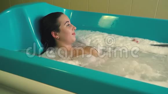 穿着泳衣的黑发女人躺在泡沫浴缸里用于水疗慢慢地看着一边视频的预览图