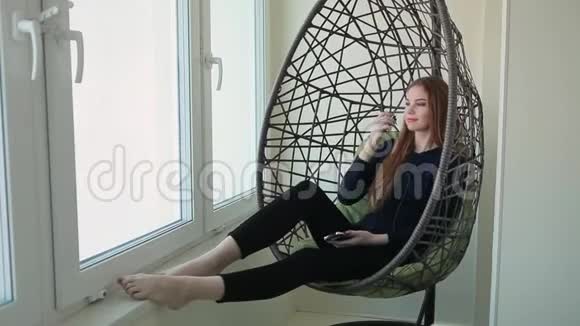 美丽的年轻女子坐在挂椅上拿着电话听音乐视频的预览图
