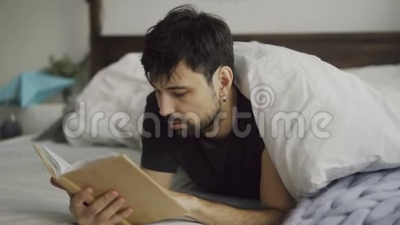英俊的年轻人在家里躺在毯子下看书视频的预览图