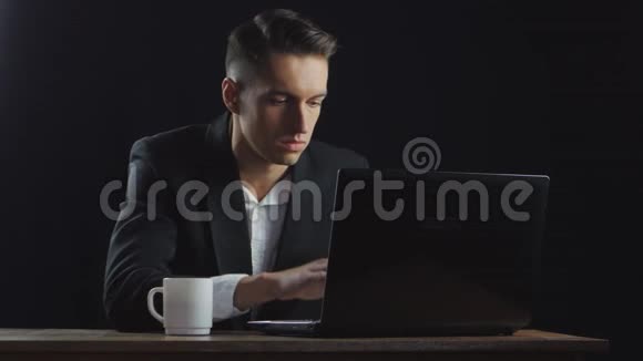 年轻的商人一边喝咖啡一边在黑暗的办公室里工作笔记本电脑视频的预览图