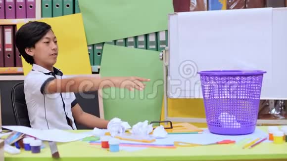 那个男孩坐在桌子上桌子上有一个废篮赌气地扔着纸团视频的预览图