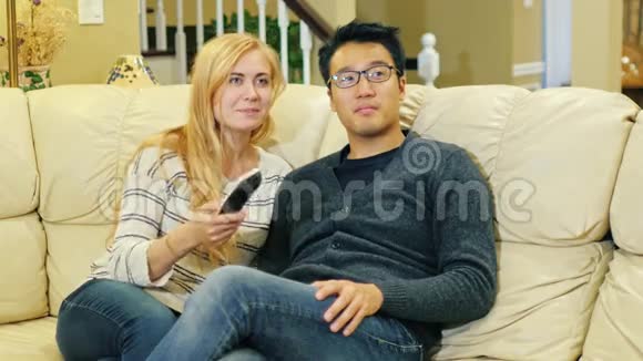 多种族夫妇在家看电视亚洲男人和高加索女人坐在沙发上手里拿着遥控器视频的预览图