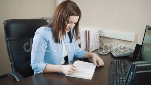 穿着蓝色夹克的黑发女商人微笑着在办公室里写日记视频的预览图