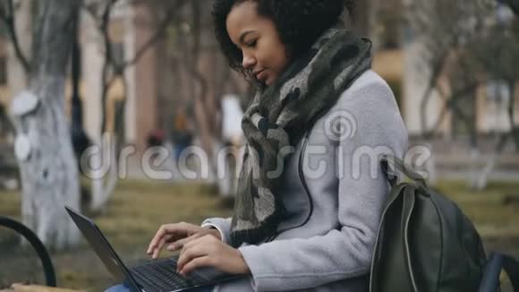 漂亮的非裔美国学生女孩坐在大学附近的长凳上打笔记本电脑视频的预览图
