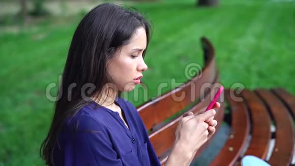 女人坐在长凳上在公园的背景上使用智能手机视频的预览图