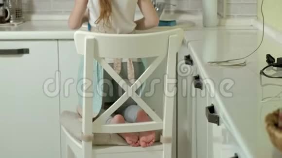穿着围裙的漂亮小女孩站在椅子上擦拭桌子明亮的厨房里的工作表面视频的预览图