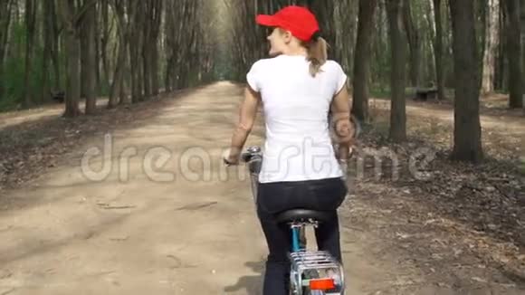 快乐活泼的年轻女子骑着红帽子骑着自行车在阳光明媚的公园夏天慢动作视频的预览图