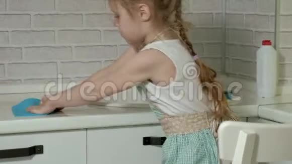 穿着围裙的漂亮小女孩站在椅子上擦拭桌子明亮的厨房里的工作表面视频的预览图