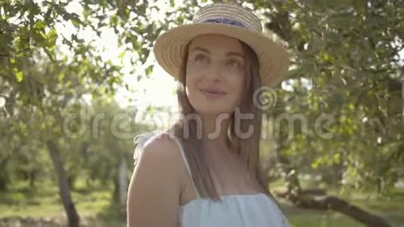 漂亮的年轻女子戴着草帽穿着白色长裙看着镜头微笑地站在绿色的夏天视频的预览图