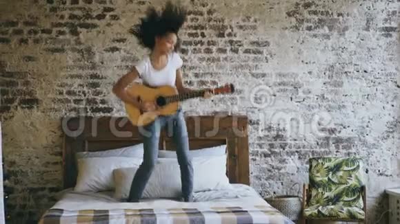 混合种族的年轻滑稽女孩弹声吉他在家里的床上尽情跳舞视频的预览图