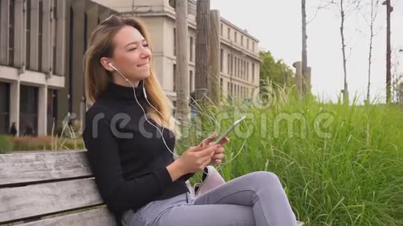 满意的女学生听音乐的白色在耳电话和平板电脑在长凳上慢动作视频的预览图