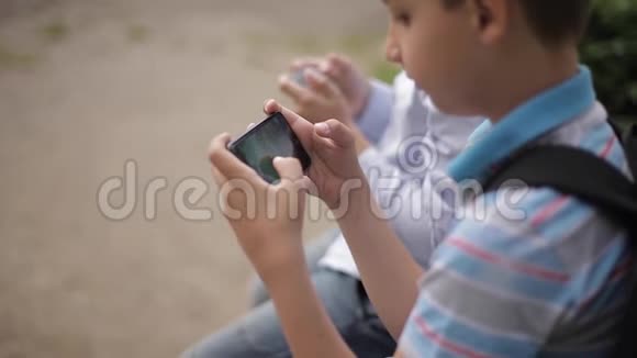 两个男孩坐在长凳上玩网络游戏一个带背包的男孩年轻人用电话视频的预览图