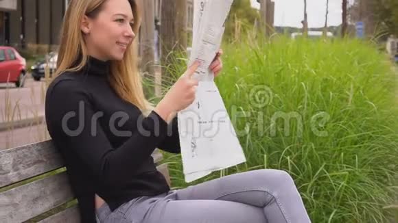 女性阅读报纸和坐在靠近道路的长凳上慢动作视频的预览图