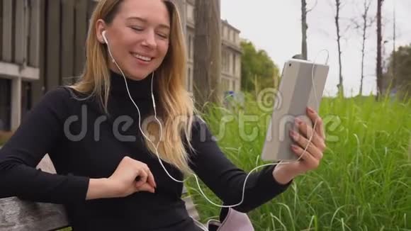 女人通过平板电脑和带视频电话的耳机说话坐在长凳上慢慢地竖起大拇指视频的预览图