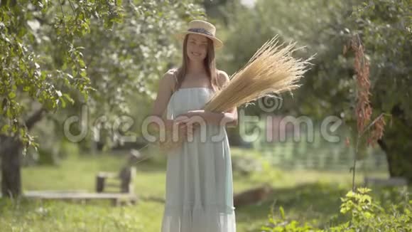 漂亮的年轻女子戴着草帽穿着白色长裙看着镜头微笑地站在绿色的夏天视频的预览图