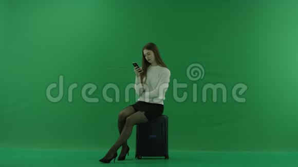女孩坐在一个手提箱上半转看着左边看着绿色屏幕上的电话视频的预览图